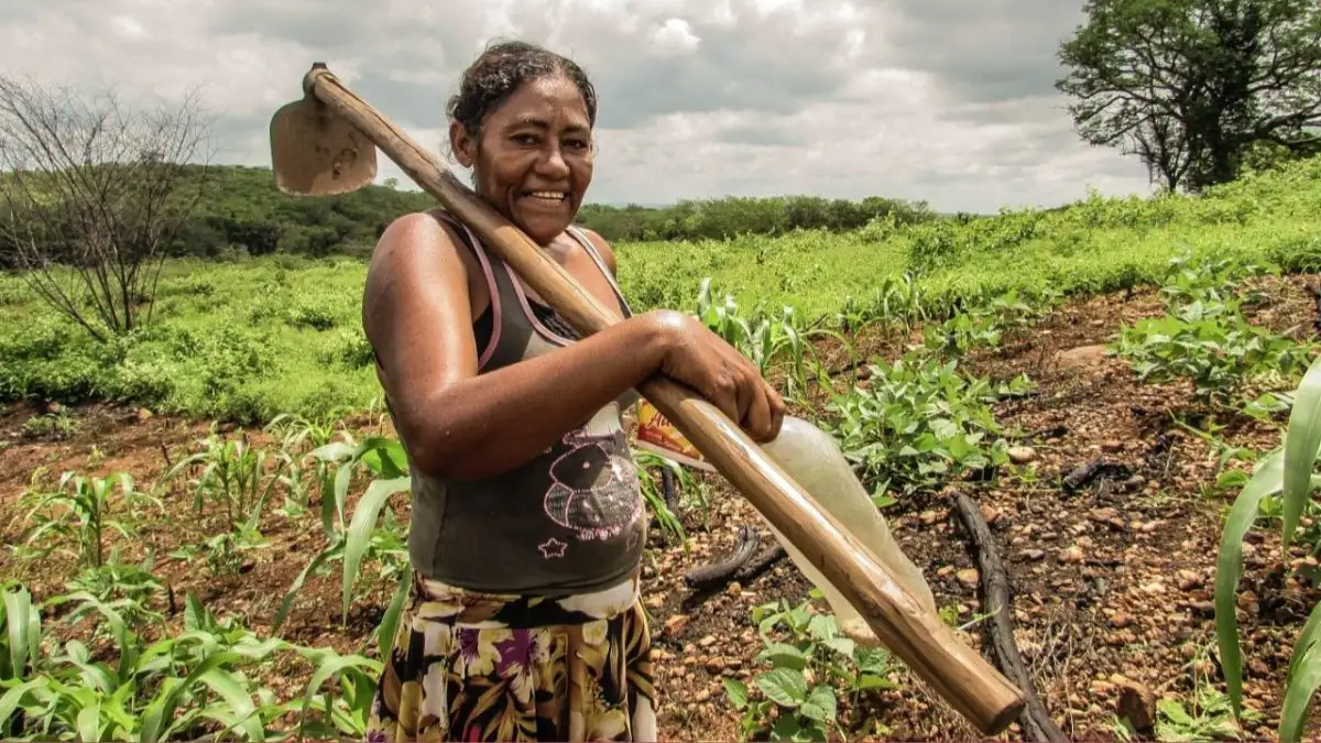 Produtora rural sorrindo, com enxada no ombro, no meio da plantação